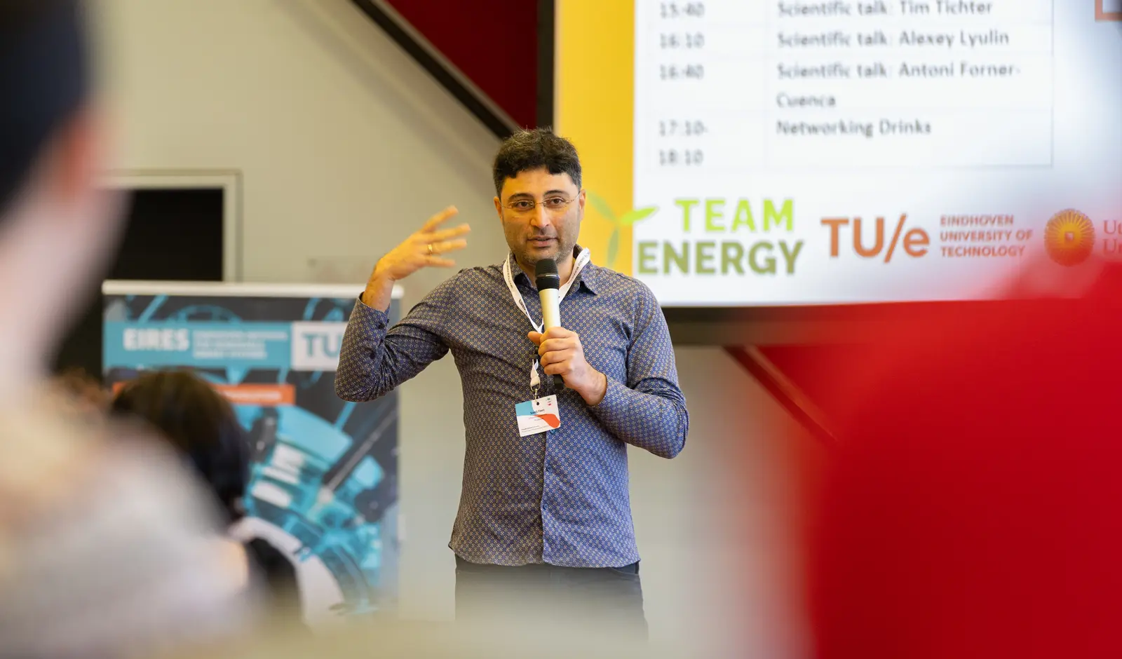 A man with a microphone in front of a projector screen addressing an audience while gesturing